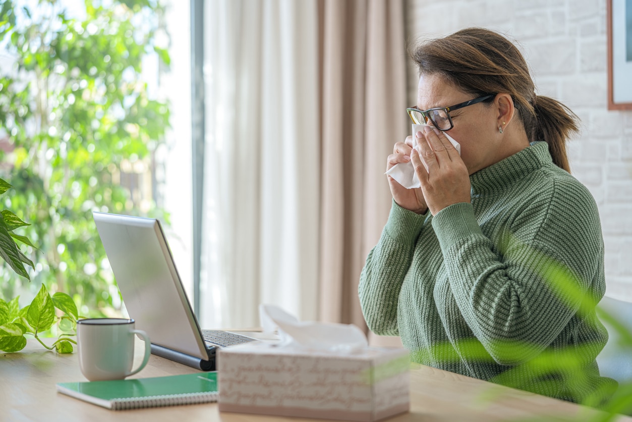 Woman at desk with laptop in front of her blowing her nose.